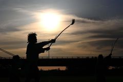 帯広カムイトウウポポ保存会による弓の舞「ク　リムセ」。弓を掲げる男性を夕日が照らす。狩人が鳥を撃とうとした時、鳥が美しく撃つことができなかったという物語からこの踊りができたと伝わる。神々への奉納の踊りとして儀式や祭事の時に踊られるほか、近年では男性の代表的な踊りとして公演などで披露されている