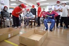 箱の中の紙皿に乗せたボールを落とさないよう、ひもで箱を引き寄せる「ひもまきまきリレー」を楽しむお年寄りら